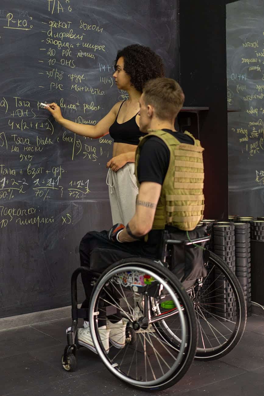 a man sitting on a wheelchair while listening to the woman writing on the board