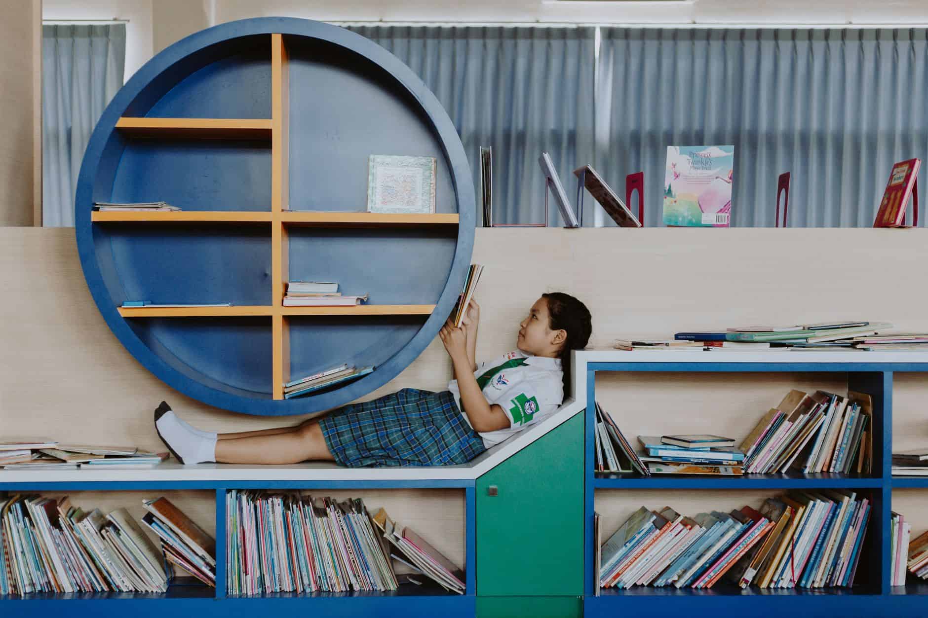 girl in school uniform lying on book shelf and reading