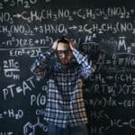 man in eyeglasses holding his head next to blackboard with math equations