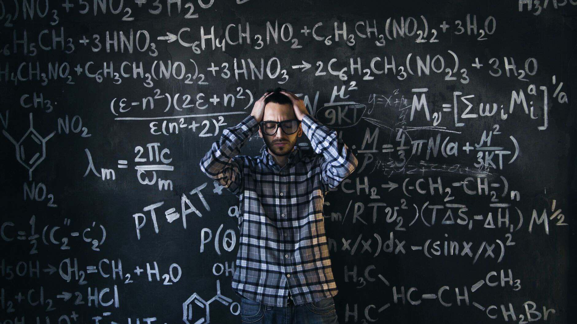 man in eyeglasses holding his head next to blackboard with math equations